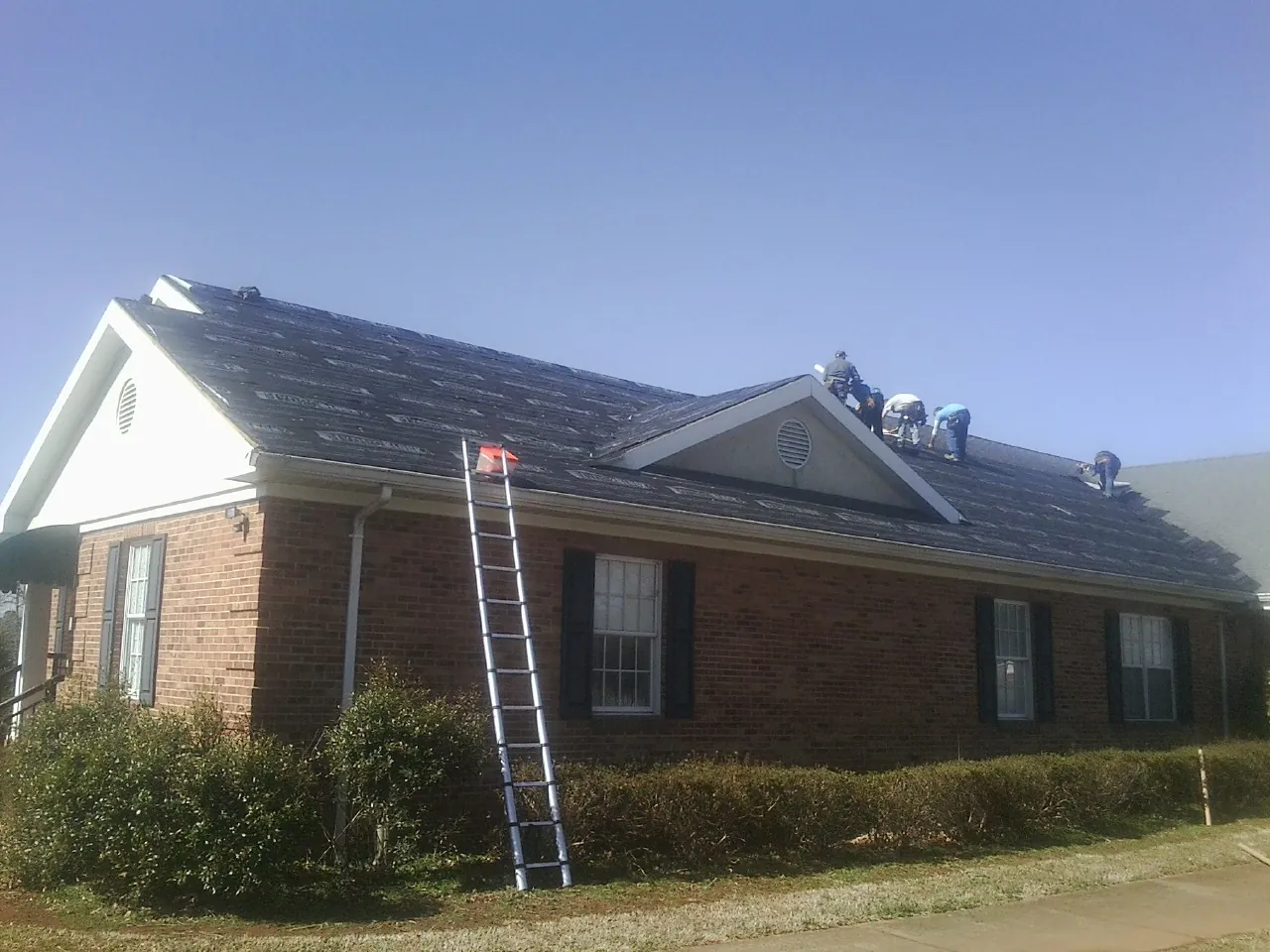 Crew tearing shingles off a brick ranch roof with a ladder set — Emergency Roof Repair work by our crew in Greenhurst