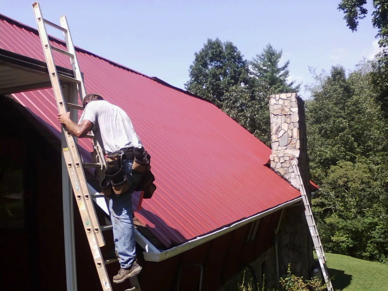 Roofer on a ladder beside a red metal roof and stone chimney — Soffit & Fascia Repair by Roofing Brothers in Greenhurst